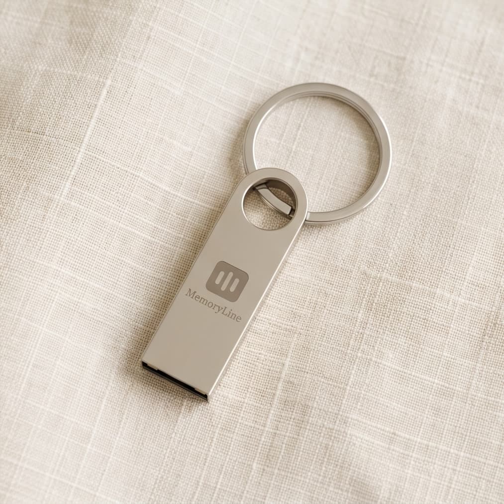 A silver keepsake USB on a keyring, the MemoryLine mark etched into the metal, on cream linen.